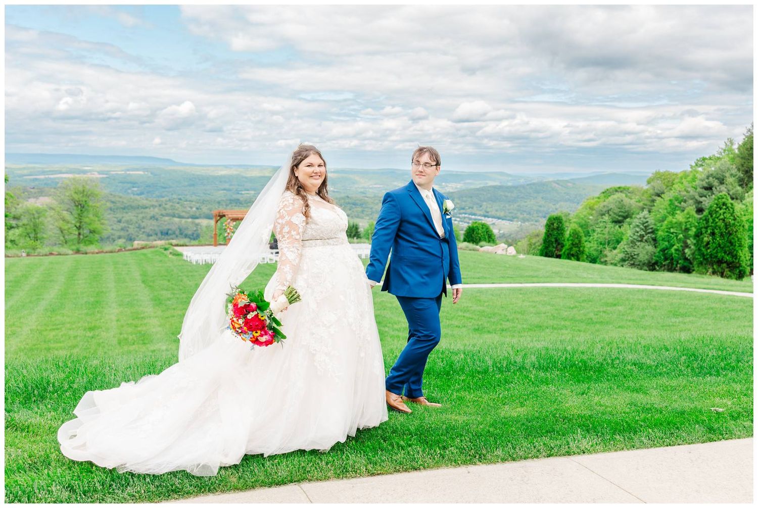 A couple walks on a lush hilltop lawn surrounded by mountains and greenery while holding a colorful bouquet.