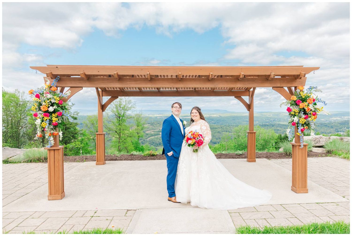 A couple stands under a wooden pergola decorated with colorful floral arrangements against a scenic mountain backdrop.
