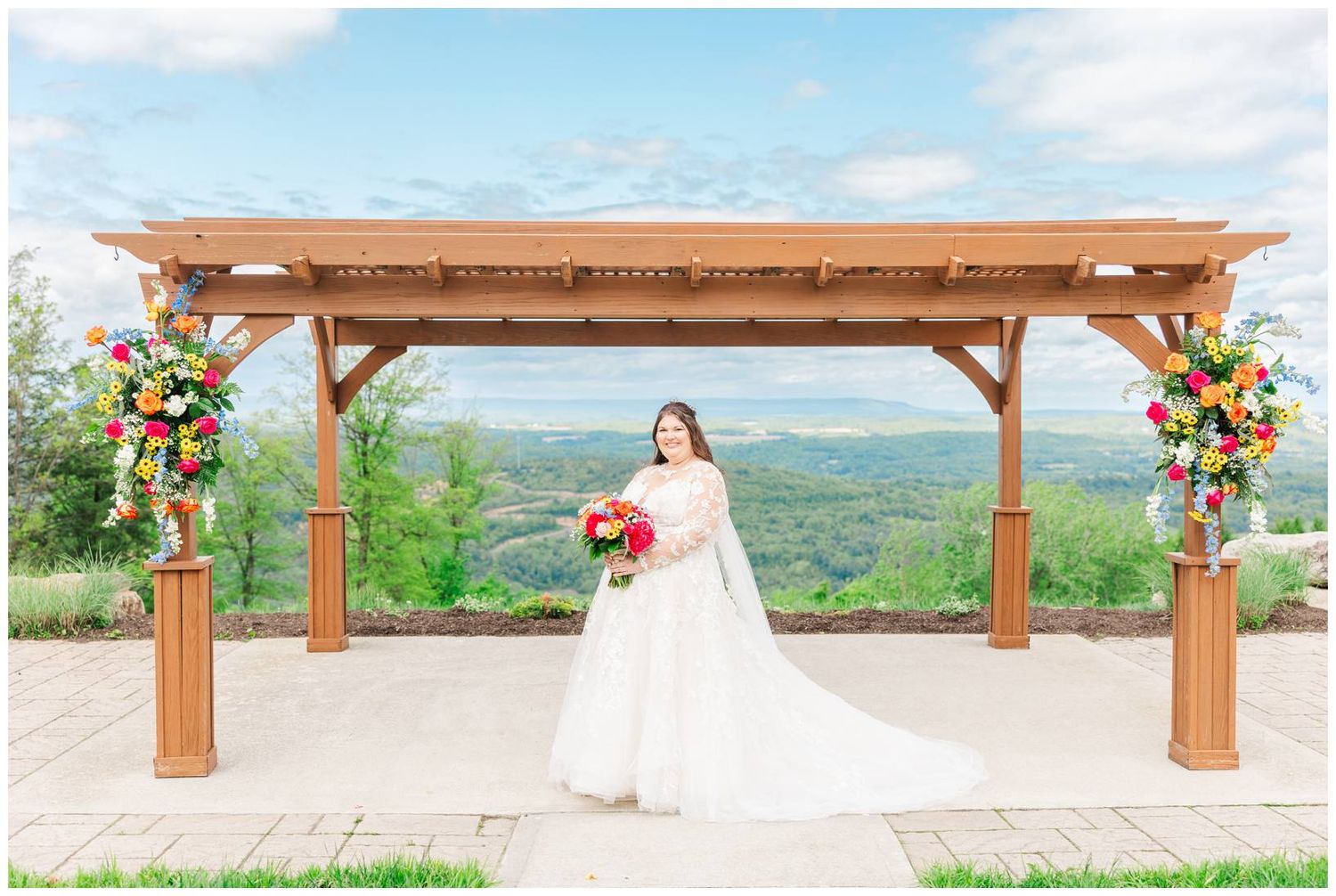 A bride in a white gown stands under a wooden pergola decorated with bright floral arrangements overlooking mountain views.