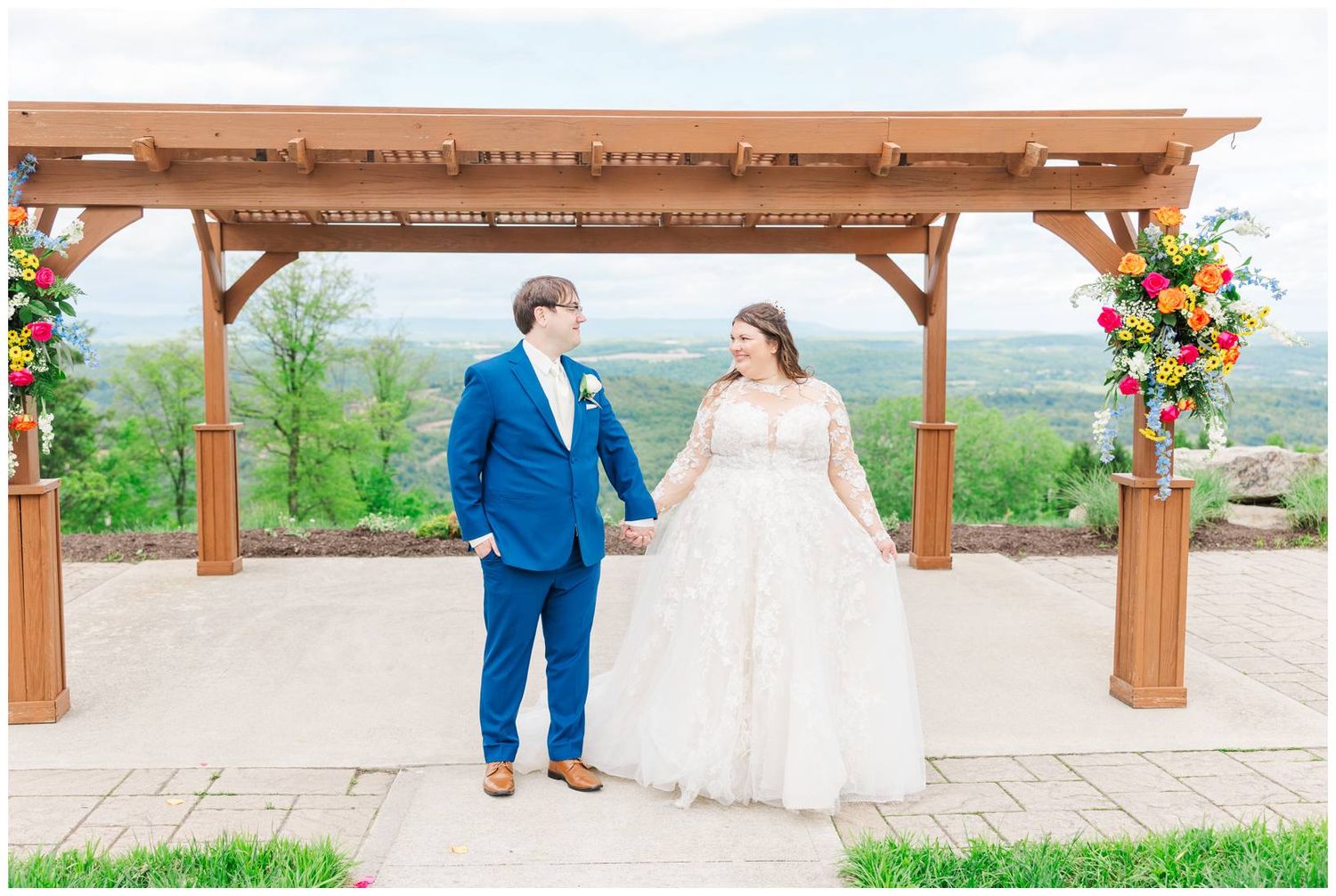 Two people standing together under a wooden pergola decorated with colorful floral arrangements in an outdoor setting.