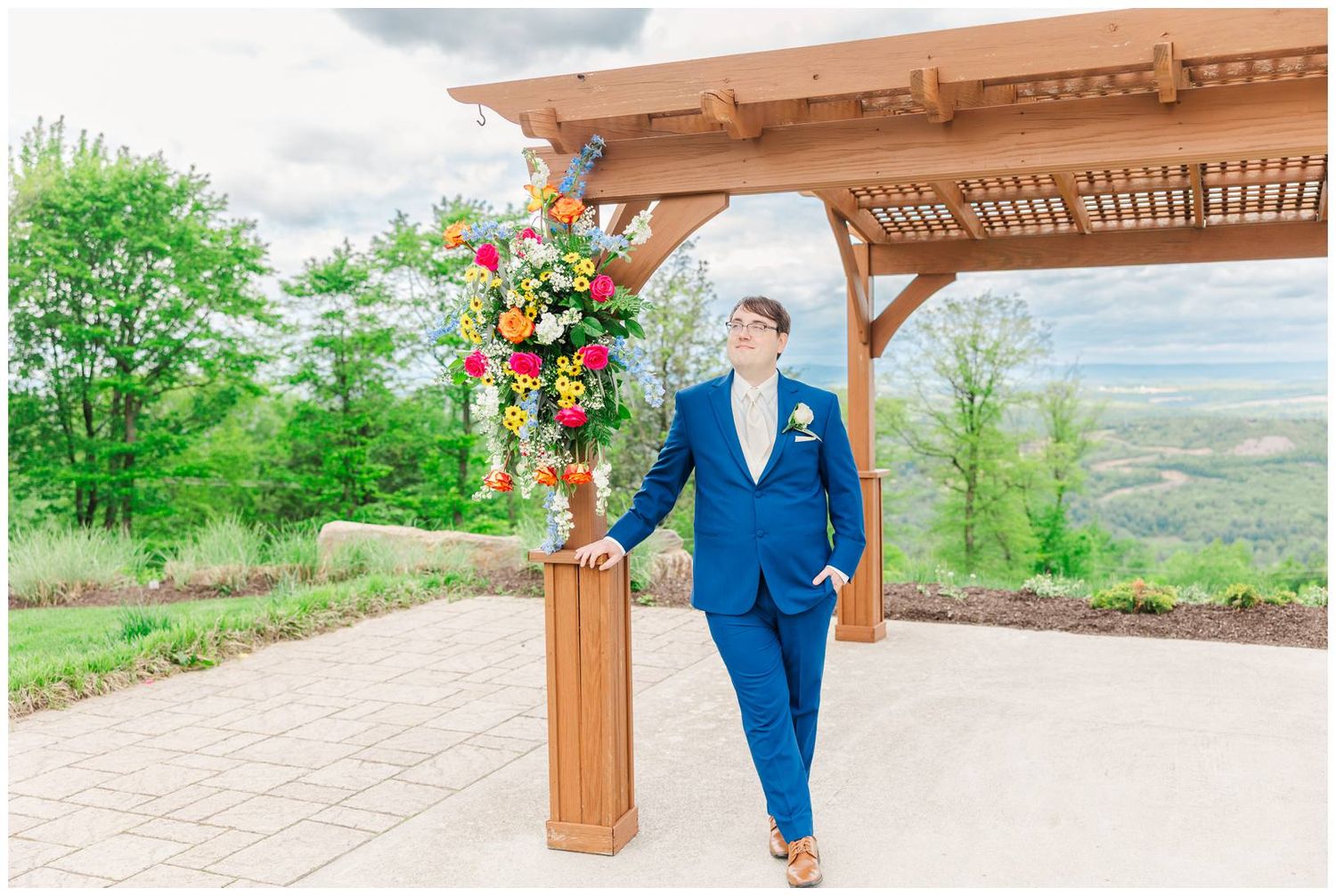A person in a blue suit stands under a pergola decorated with colorful floral arrangements on a hilltop terrace.