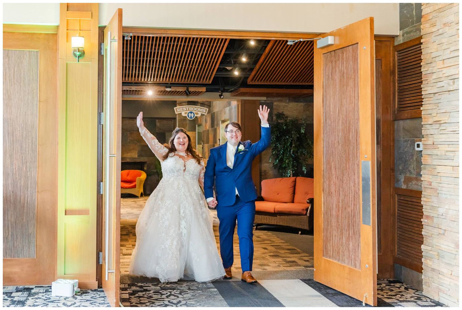 Two happy people walk through open wooden doors while raising their arms in celebration wearing formal attire.