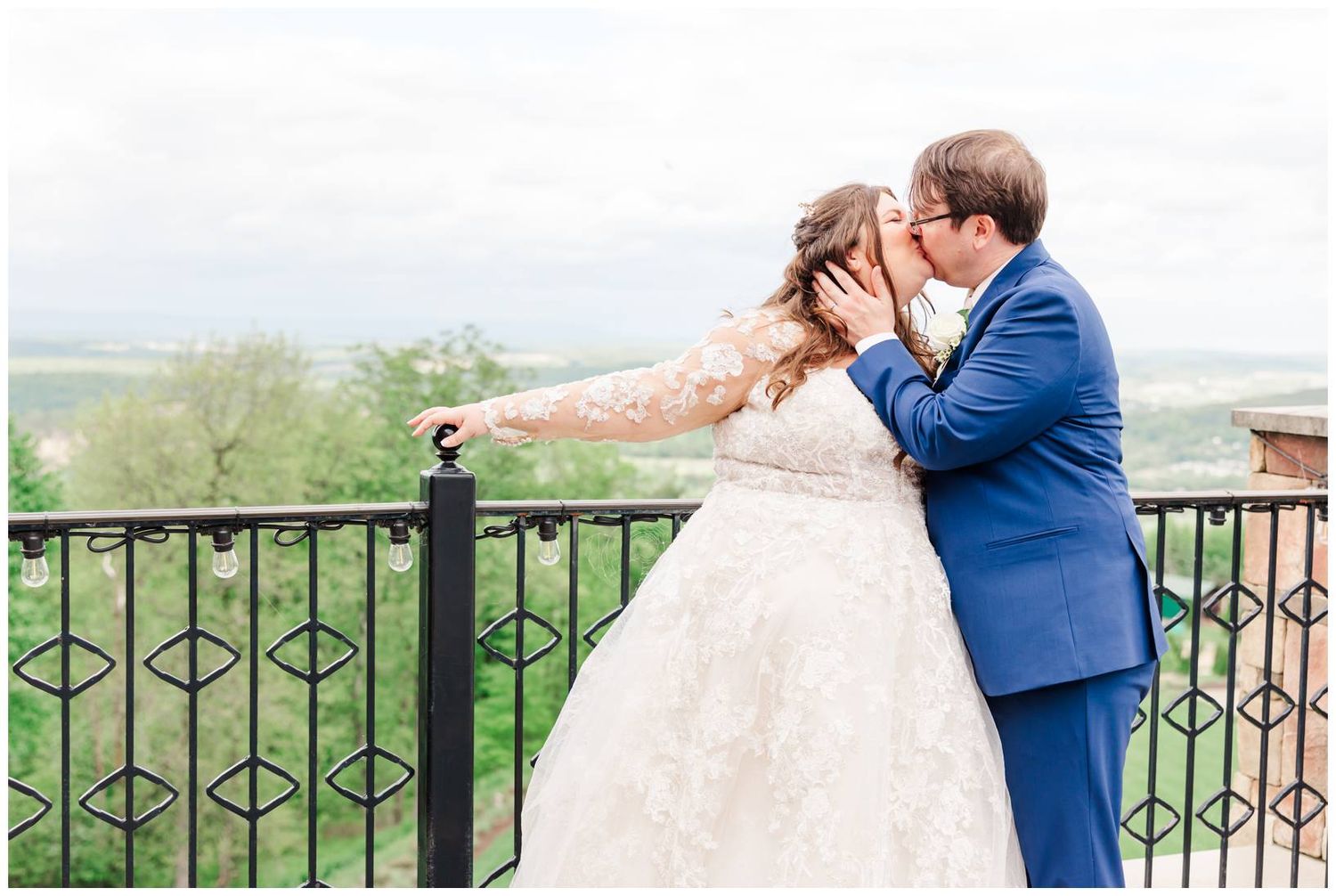 Couple in wedding attire share romantic moment on balcony with decorative iron railing overlooking scenic landscape.