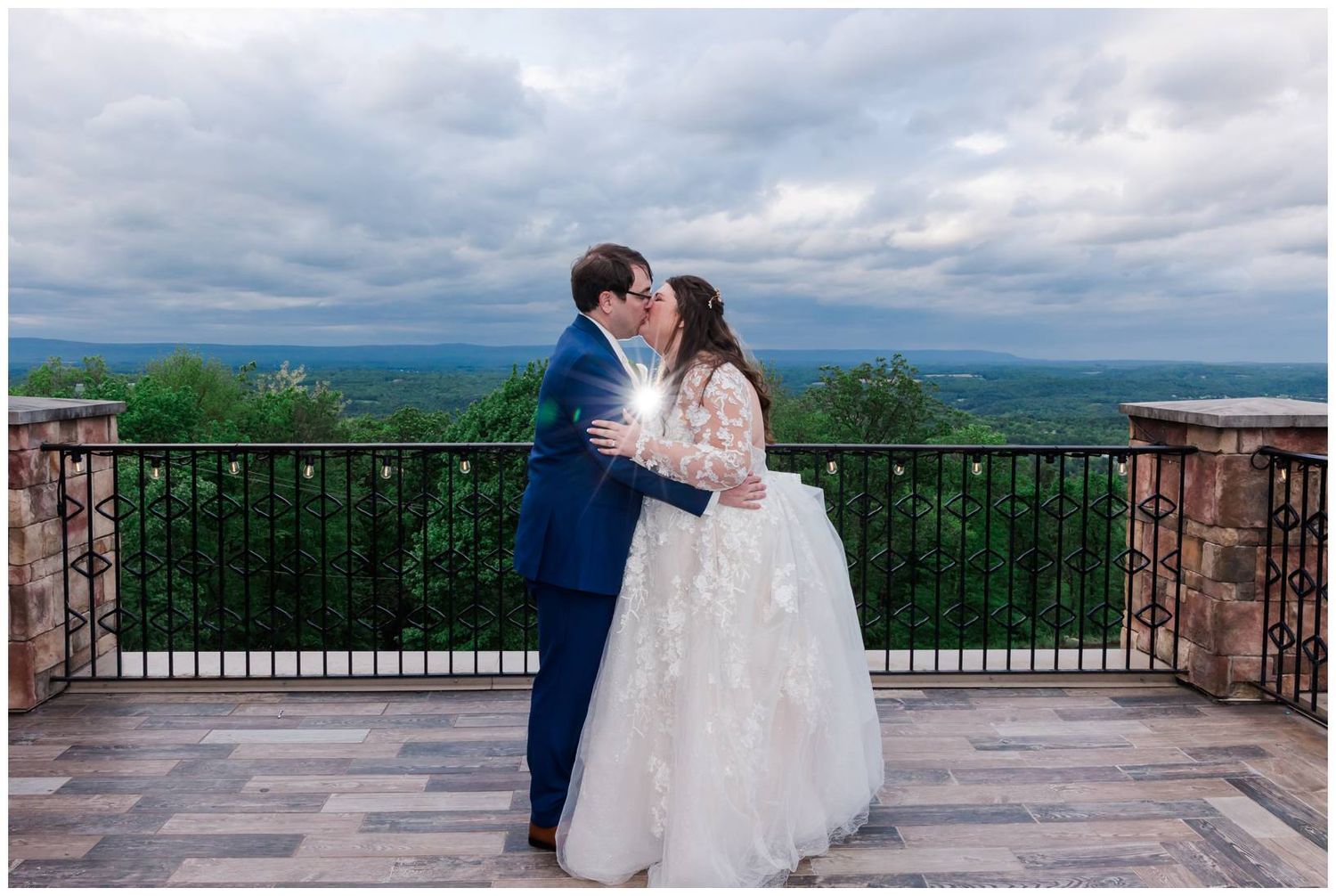 A romantic wedding couple embraces on a stone balcony terrace overlooking a scenic mountain landscape under cloudy skies.