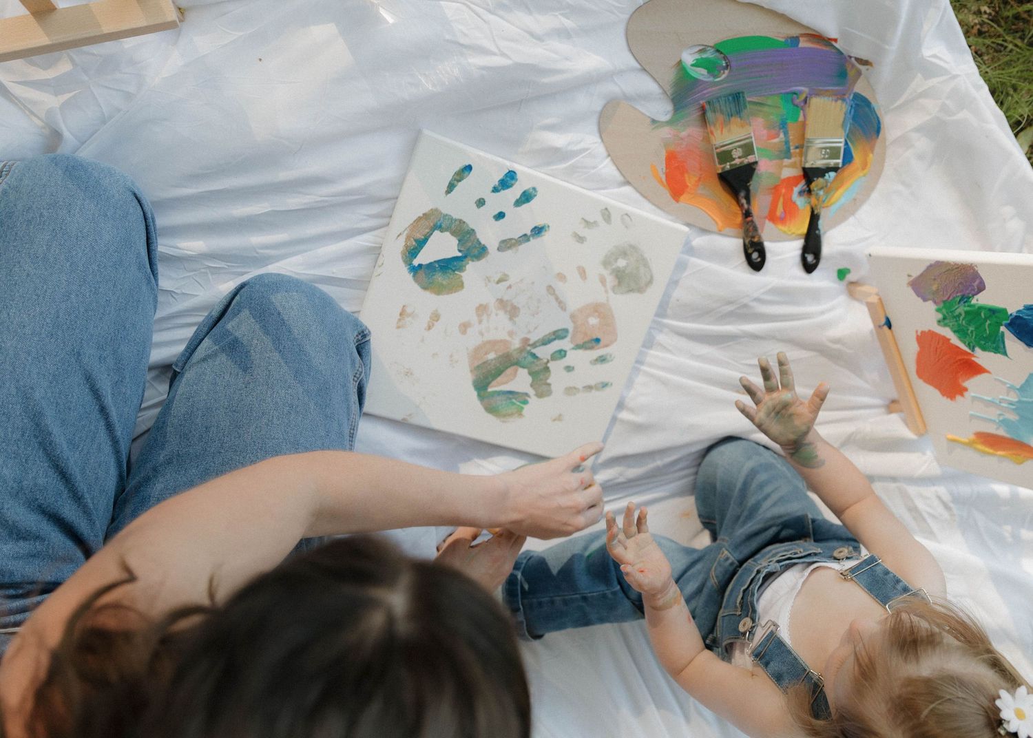 Children doing hand painting art projects outdoors on white canvas with colorful paints and green designs.