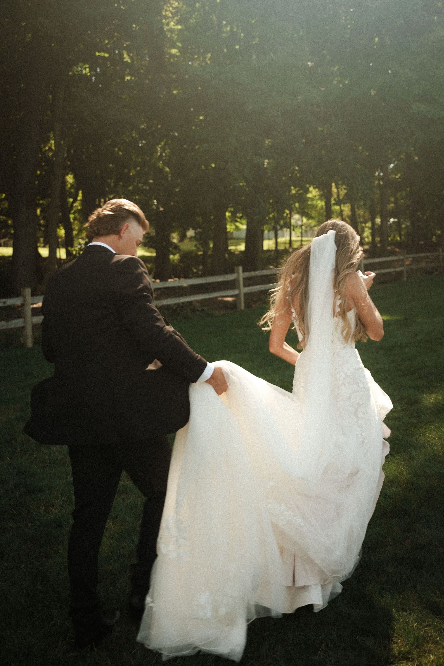 Couple walking through park with flowing white dress in golden sunlight.