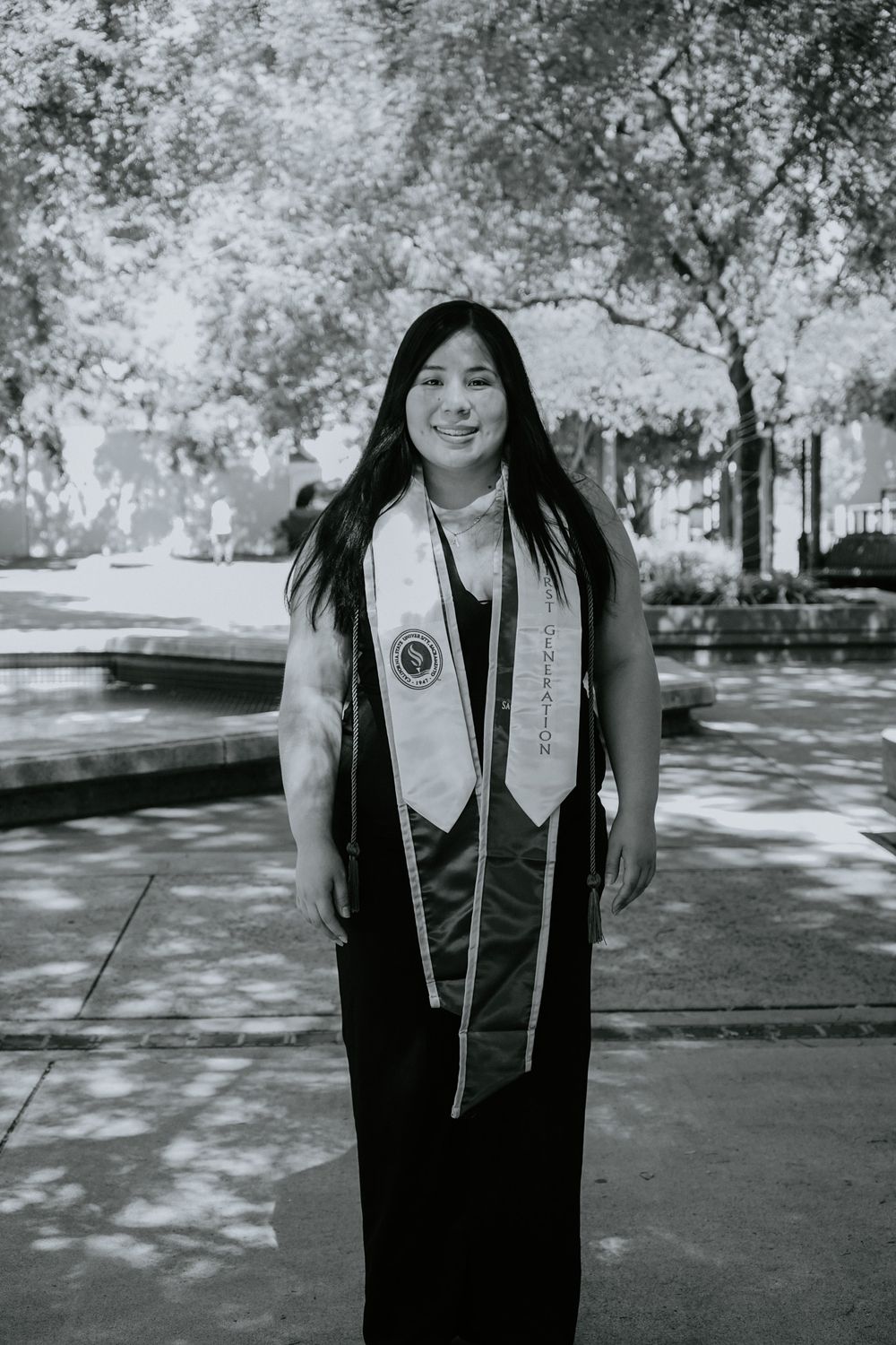A graduate in black regalia and graduation stoles stands on a tree-lined pathway in black and white photography.