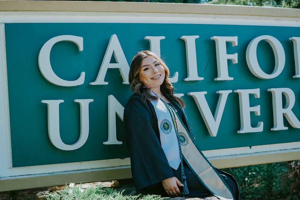 Graduate in academic regalia poses in front of a California University sign on campus.