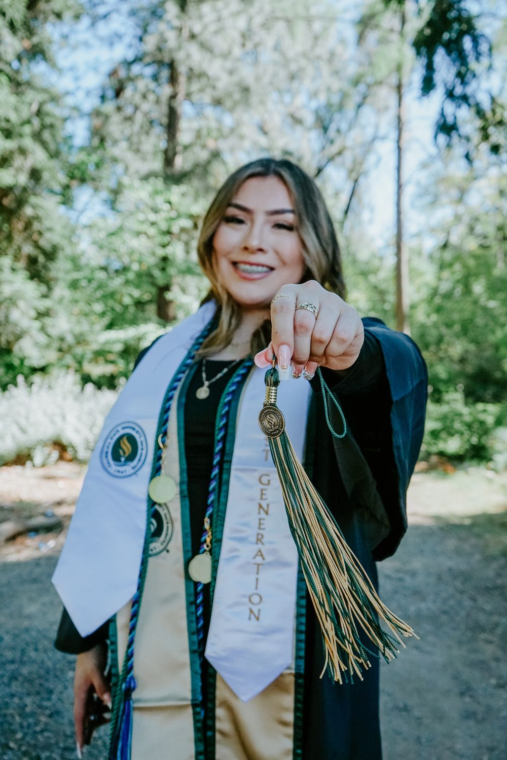 Graduate wearing graduation stole and cords displays tassel in outdoor photoshoot with trees in background.