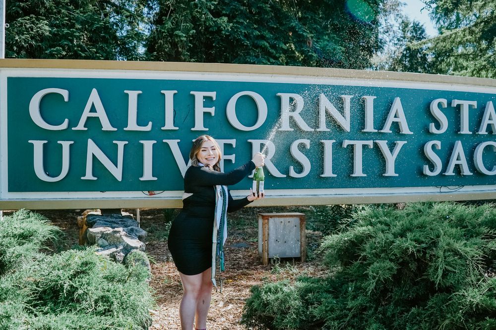 A graduate stands next to the California State University Sacramento entrance sign surrounded by green foliage.