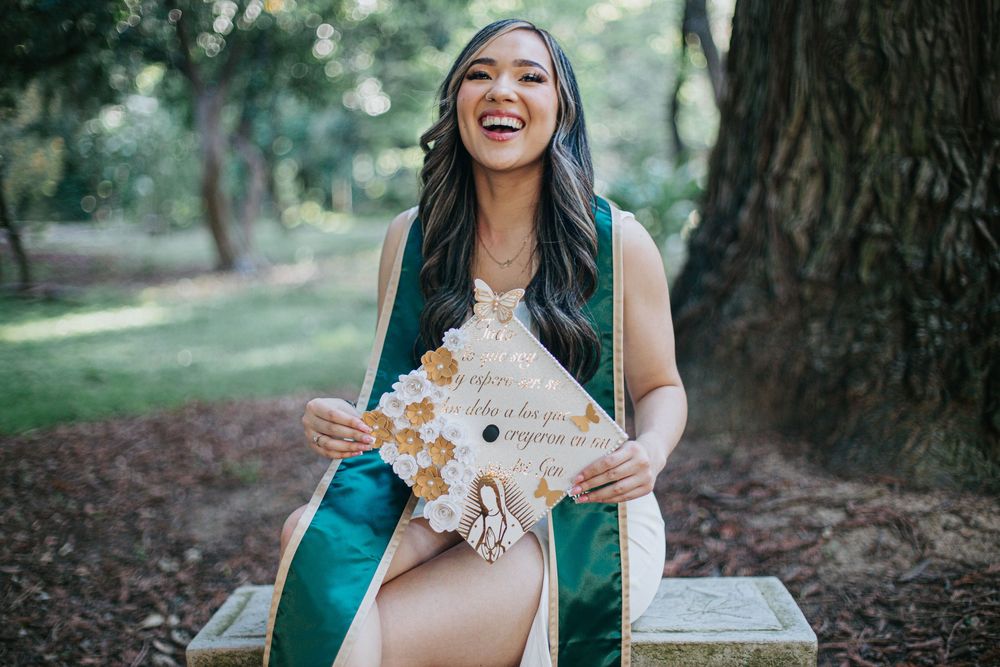 Graduate in emerald green stole and white dress joyfully poses with graduation cap on grassy lawn near tree.