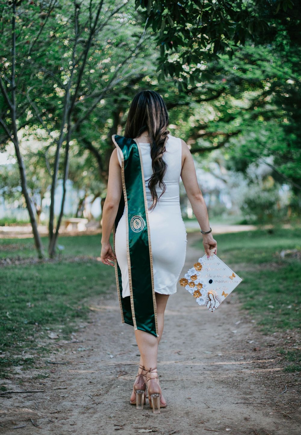Graduate wearing white dress and green graduation sash walks down dirt path in park with diploma in hand.