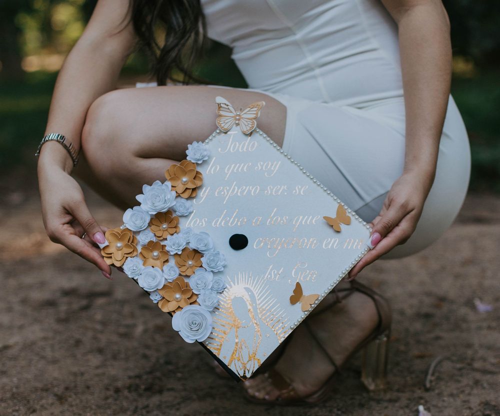 A decorated graduation cap with white and gold flowers and a bow sits on someone's lap in a white dress.
