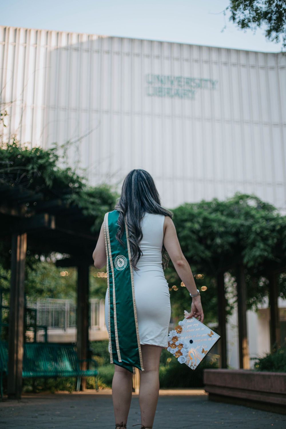 Graduate in white dress and green sash stands holding decorated cap on campus walkway.