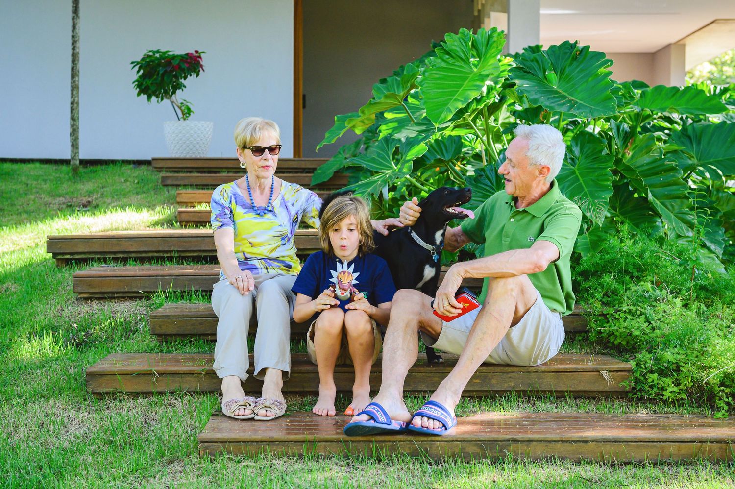 People sitting on wooden steps outdoors with lush green foliage in background on a sunny summer day.