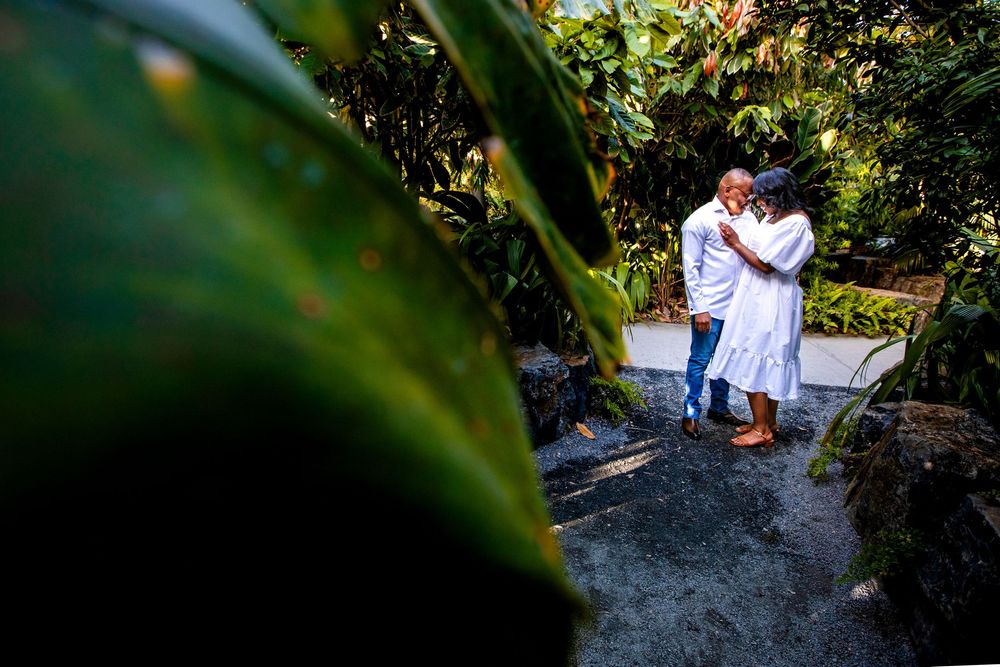 Two people in white outfits share a romantic moment in a lush tropical garden setting.