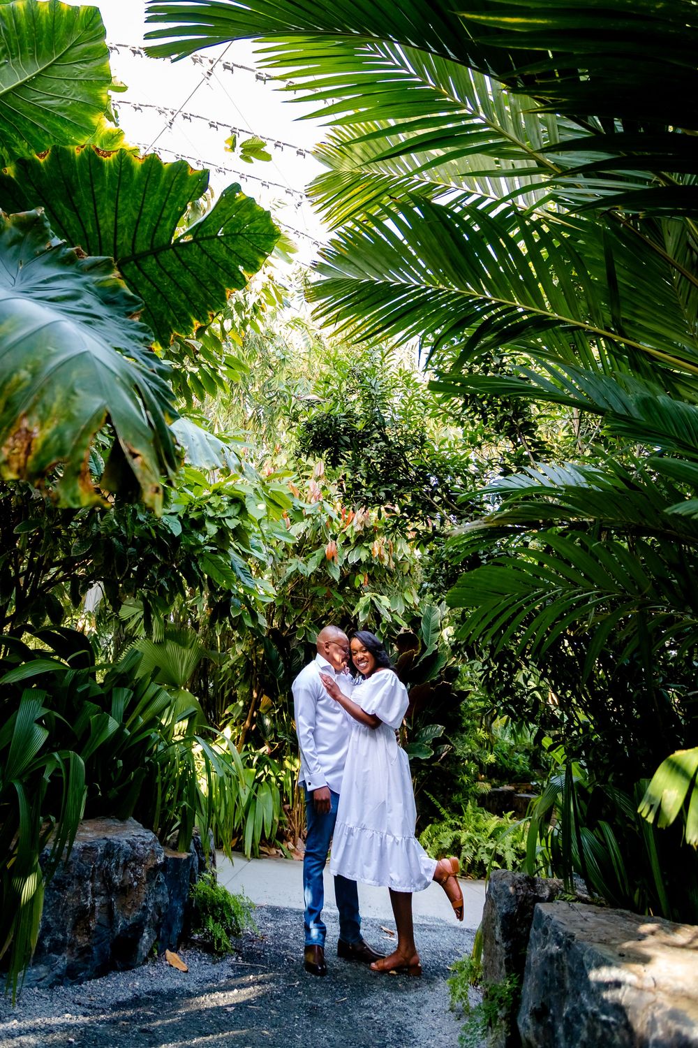 A loving couple embraces under lush tropical palm fronds in a garden pathway.