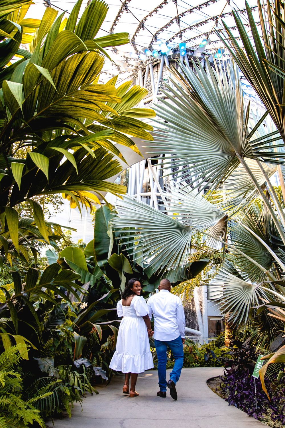Couple walking along a lush tropical garden path lined with palm fronds and string lights overhead.
