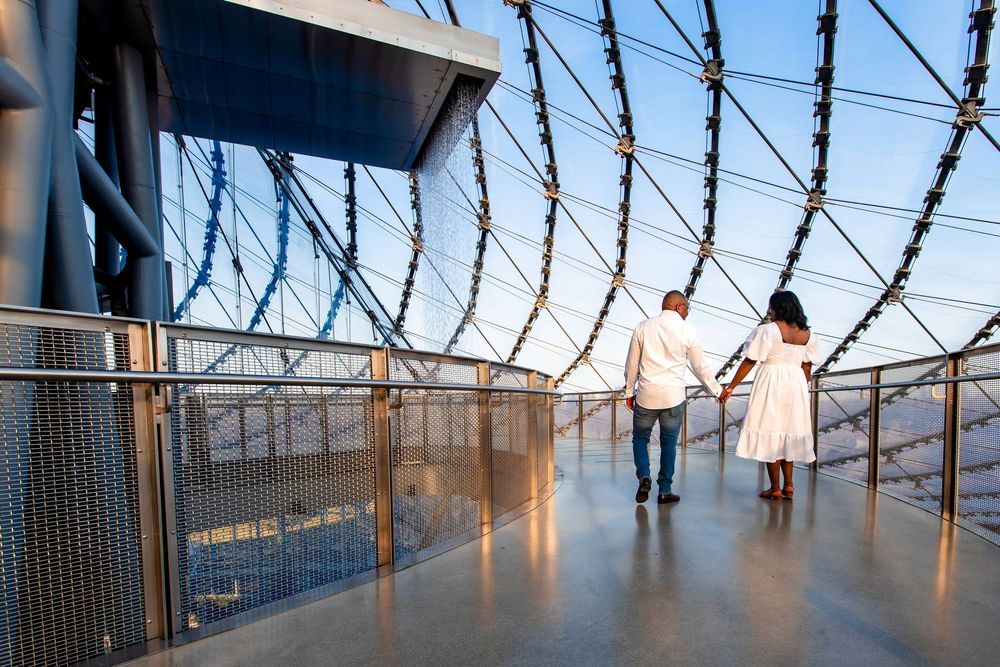 A couple walks along a curved observation deck with spiral barbed wire visible above them.