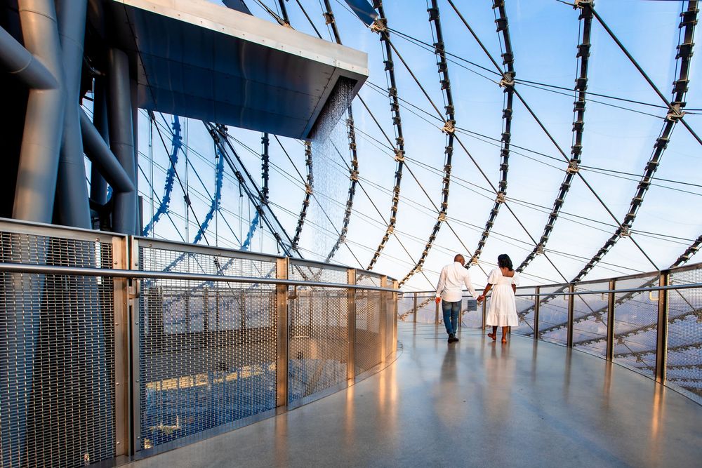Two people walk along a modern observation deck with curved glass walls and razor wire barriers overlooking a cityscape.