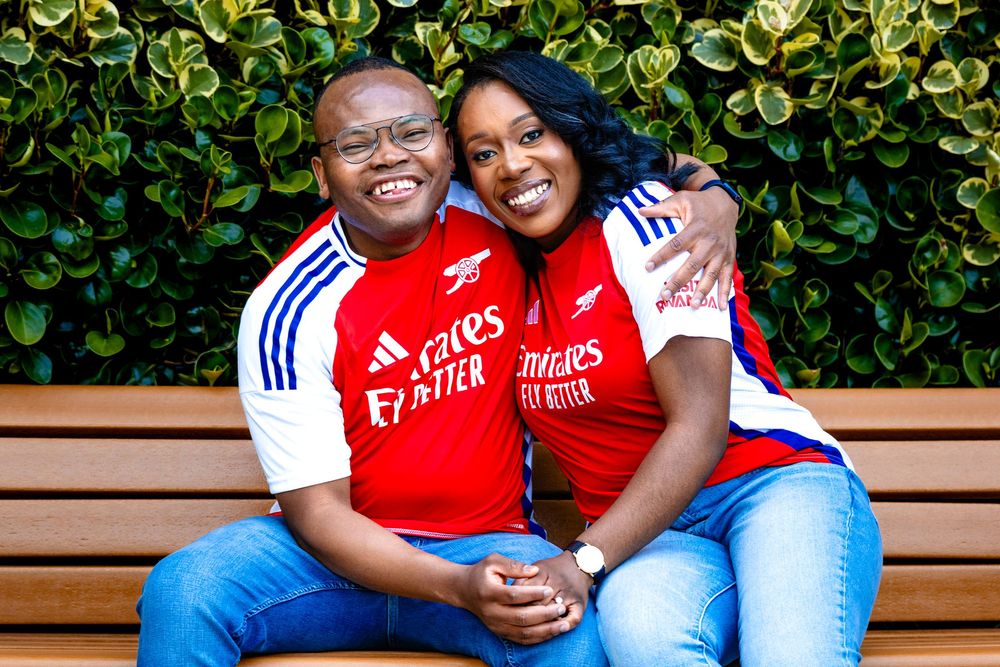 A couple in matching Arsenal jerseys sit together on a wooden bench in front of green foliage.