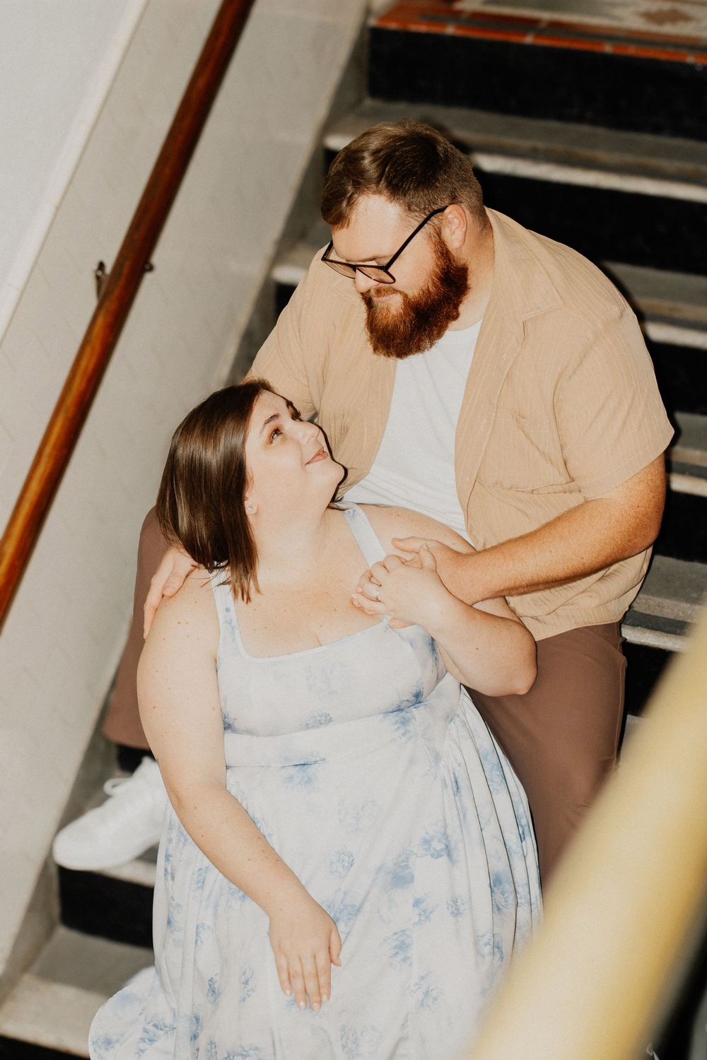 Couple sharing tender moment on staircase, woman in floral dress looking up at bearded partner.