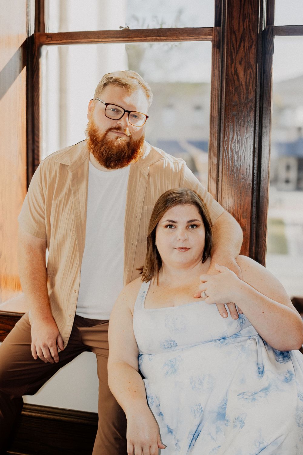 Couple posing by large window, bearded man standing behind woman seated in floral sundress.