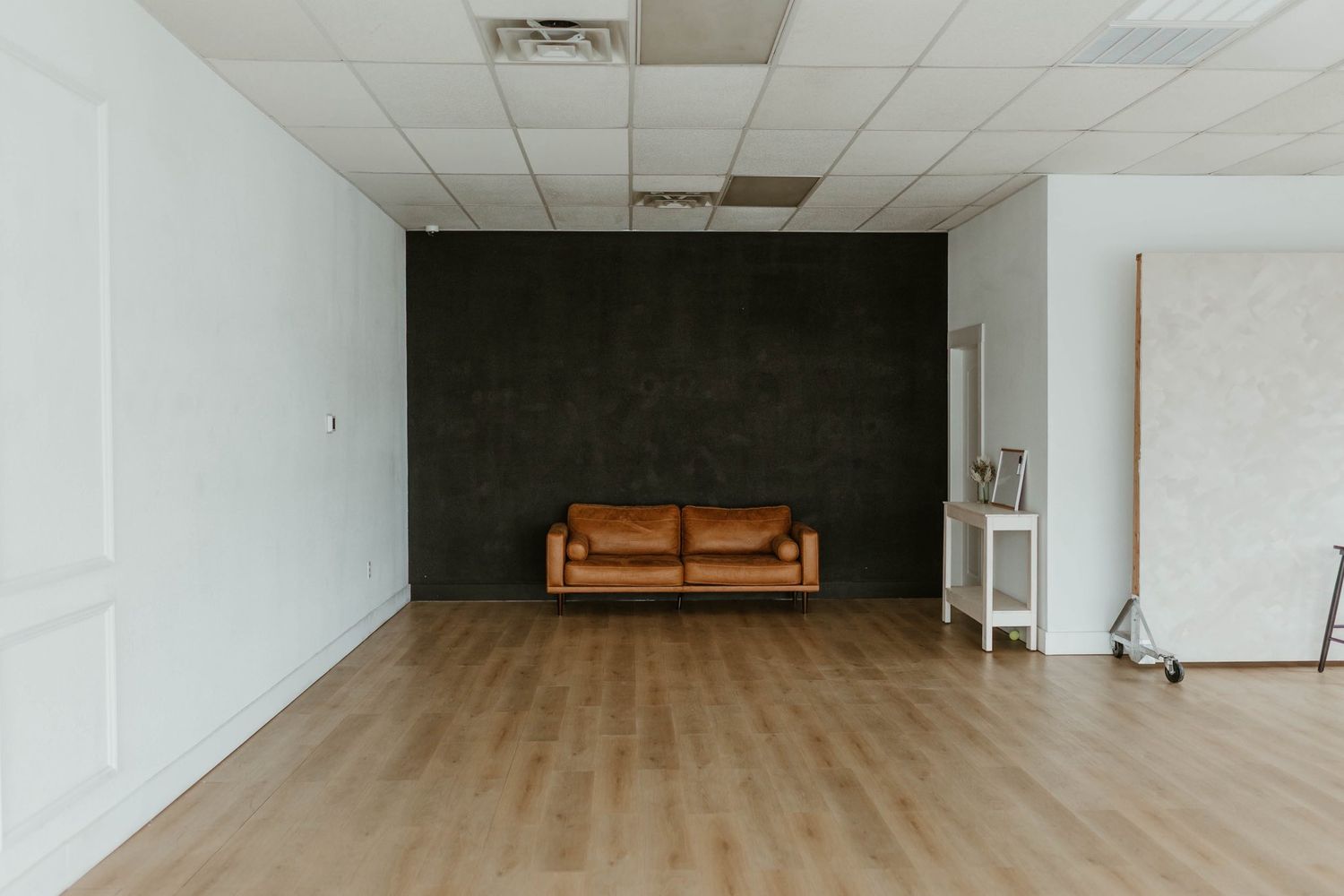 Minimalist studio space with light wood floors, white walls, black accent wall, and a lone brown leather couch.