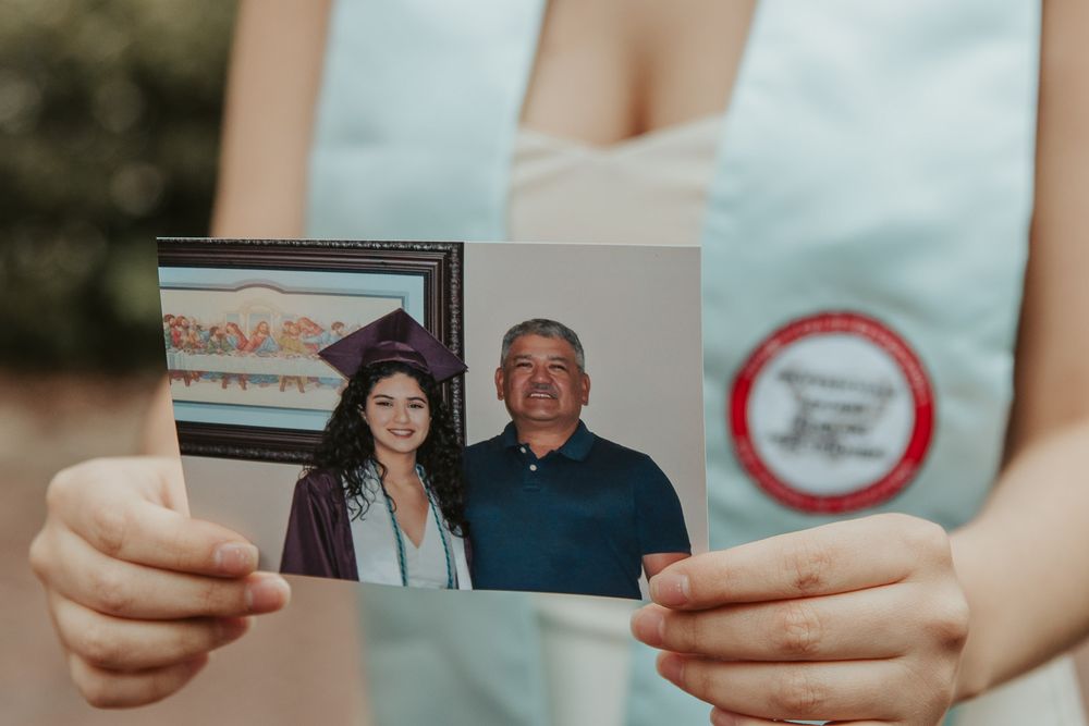 Laura smiles in graduation cap with historic Eastern Washington University Showalter Hall in the background. Holding photo of her late dad