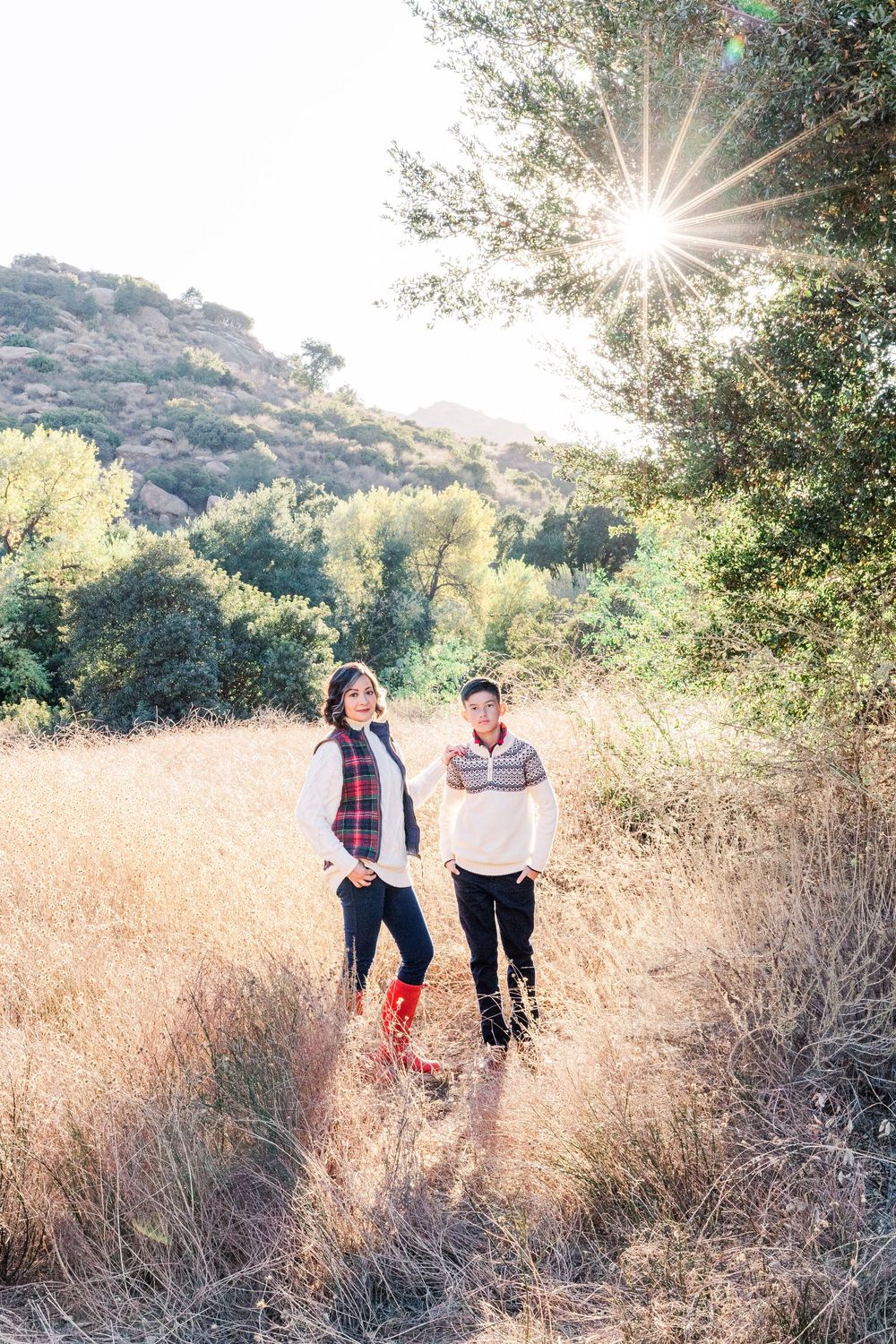 Two people walk through golden tall grass in a sun-drenched meadow with mountains and trees in the background.