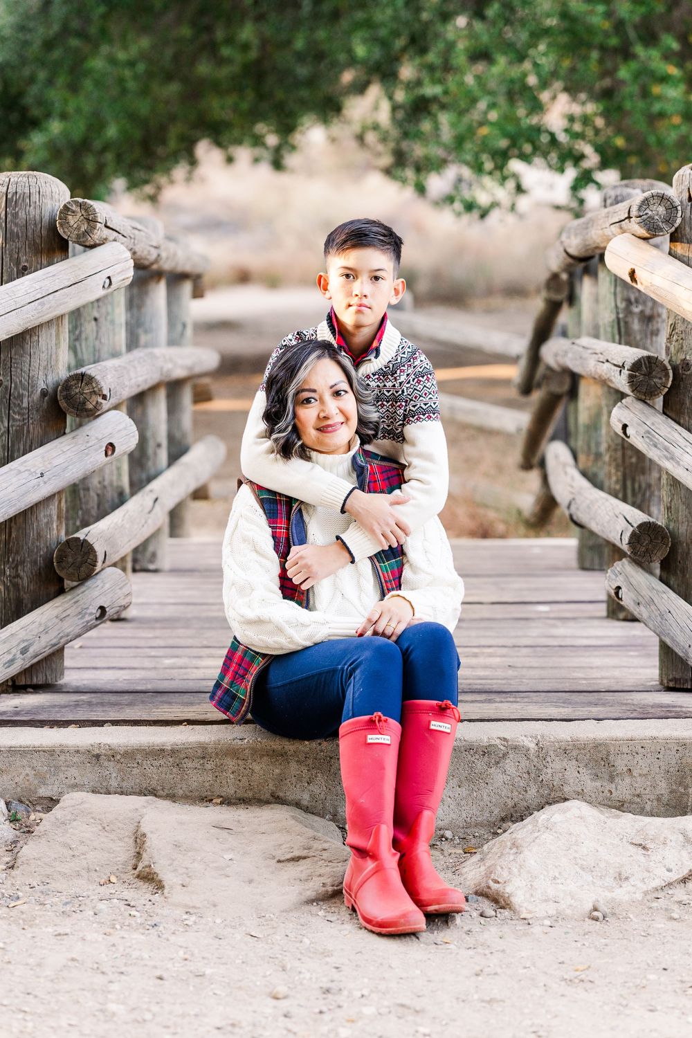 Two people sitting together on wooden bridge walkway wearing winter sweaters and red rain boots in outdoor setting.