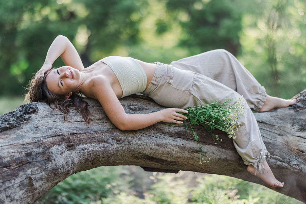 A person in beige linen clothing lies peacefully on a fallen tree trunk while holding wildflowers in a forest setting.