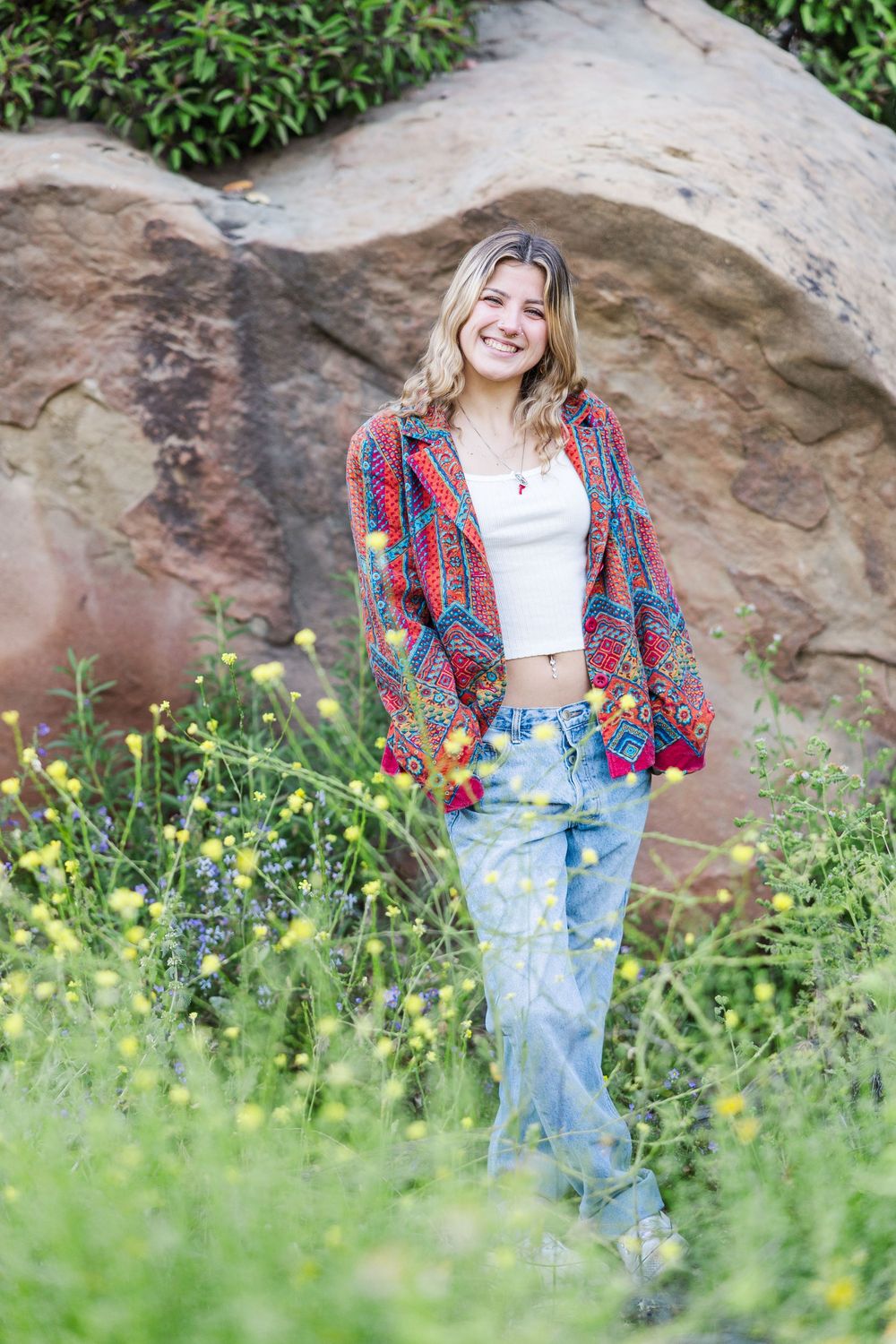 A person in a plaid shirt and jeans poses outdoors among wildflowers and rocks in a natural setting.