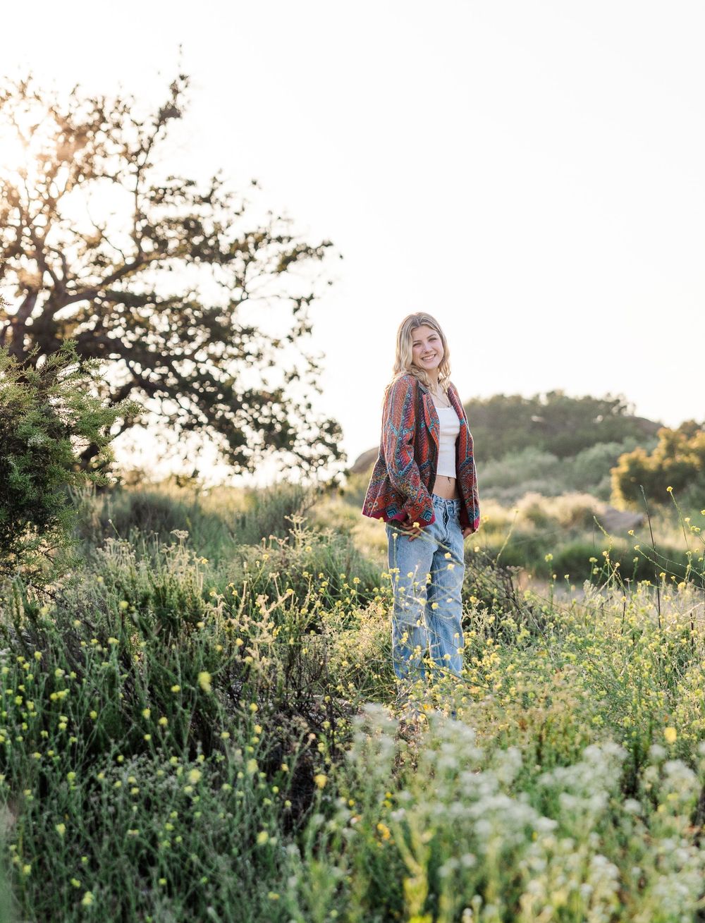 A person wearing a patterned cardigan and jeans stands in a wildflower meadow at golden hour.