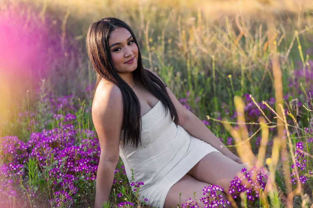 Woman in white dress poses among vibrant purple wildflowers in a sunlit meadow during golden hour.