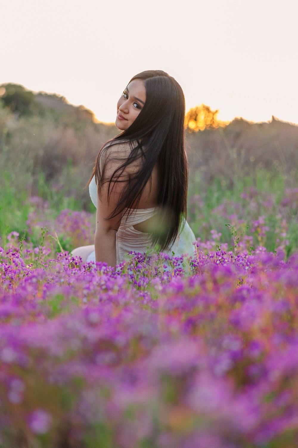 A person in a white top poses among vibrant purple wildflowers during golden hour sunset.
