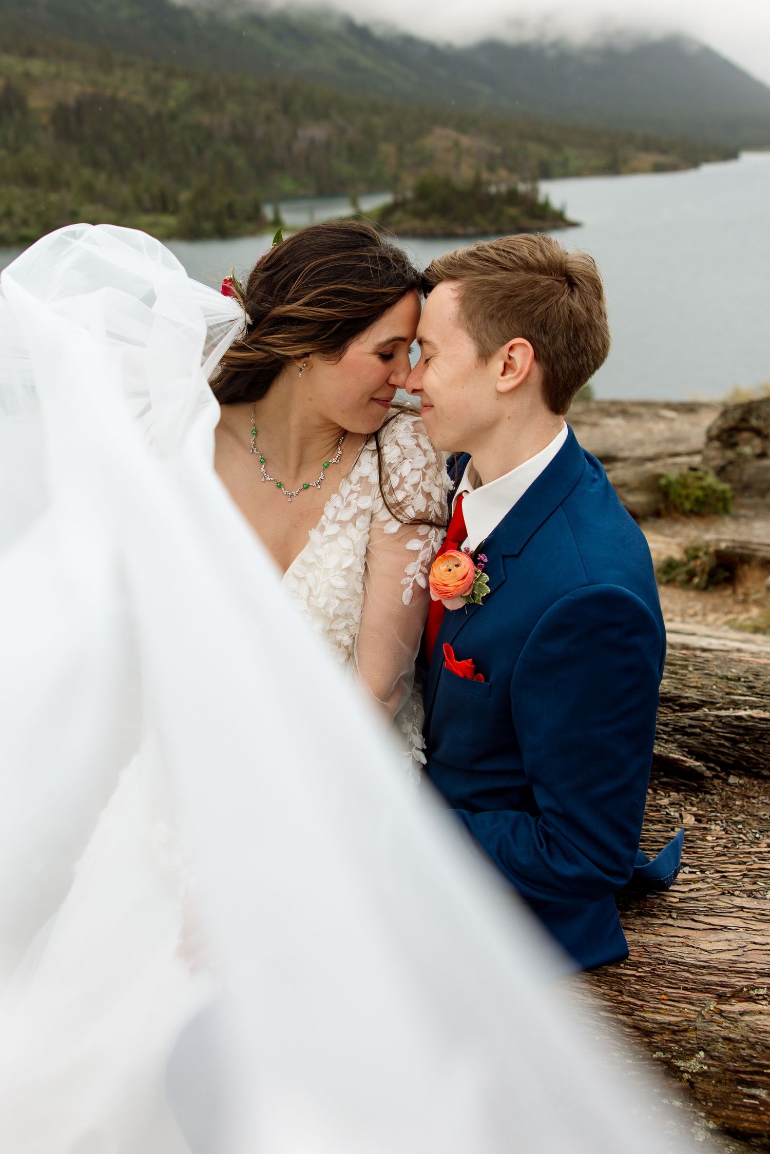 A romantic riverside wedding photo features a couple embracing by the water with mountains and pine trees behind them.