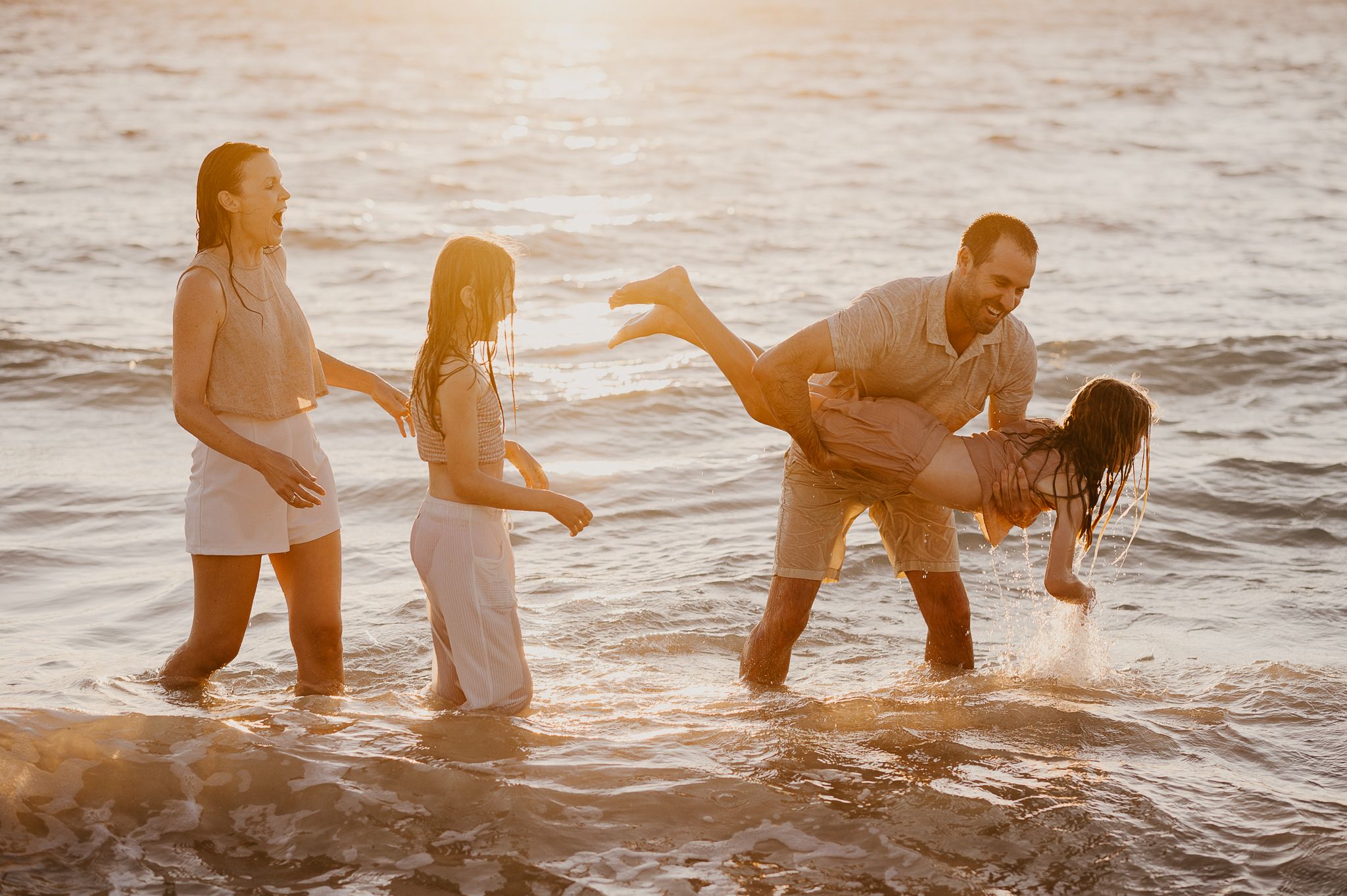 Family playing joyfully in ocean waves at sunset, parents and children laughing on a golden beach during family photoshoot Mandurah WA