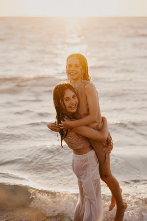 Two smiling sisters enjoy a piggyback ride on a sunny beach at golden hour for family photographer Story & Soul Photography in Mandurah WA