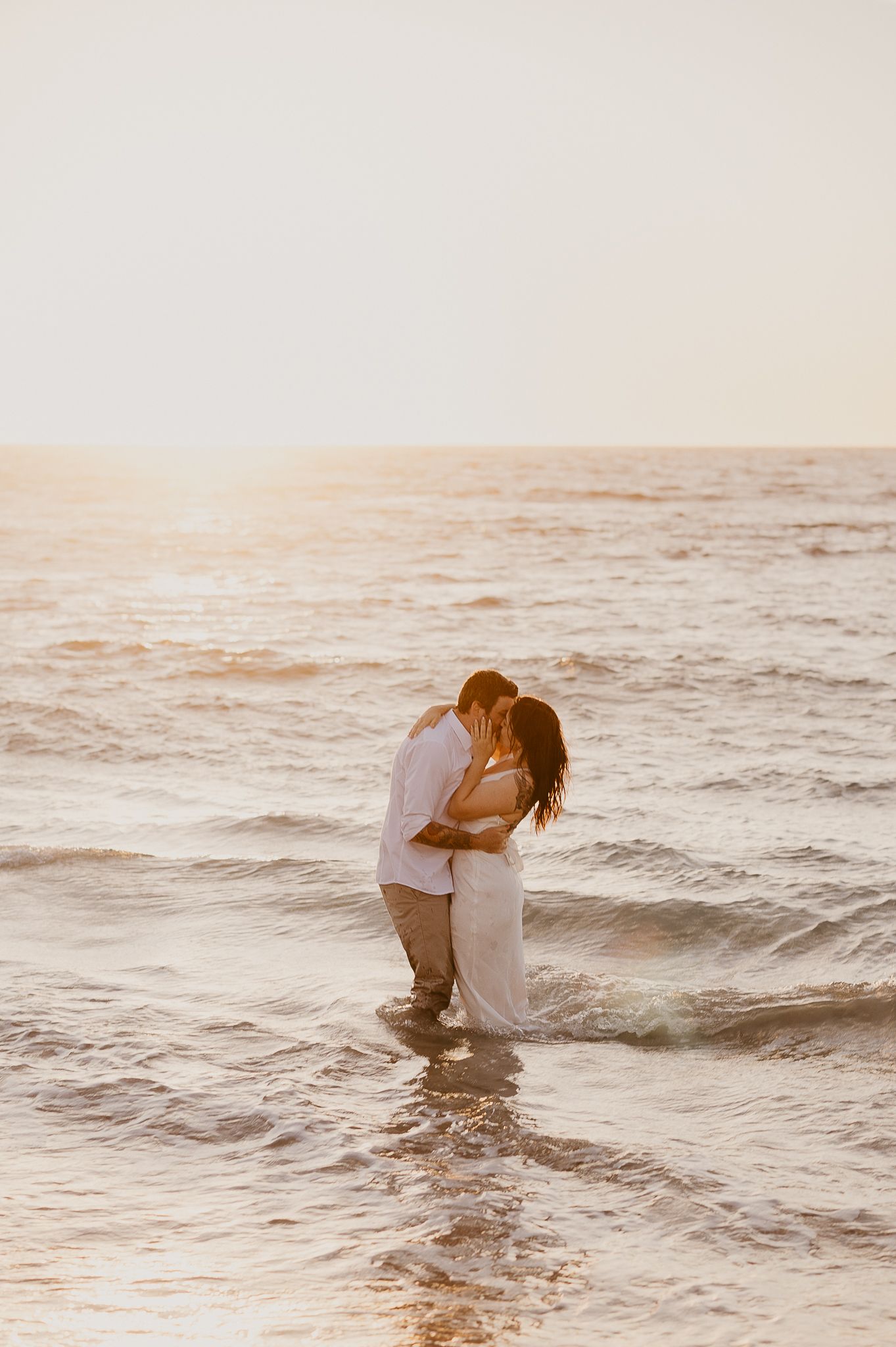 Couple kissing in ocean waves at sunset, wearing white outfits, romantic beach engagement photo with golden sun story & soul photography WA