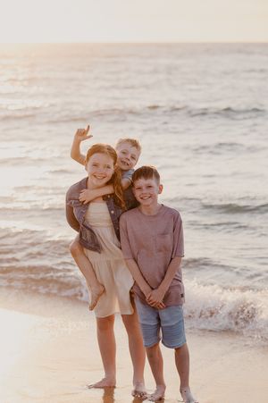 Three siblings stand on beach, waves crashing and golden sun setting for family photoshoot Mandurah. Story & Soul Photography Mandurah WA