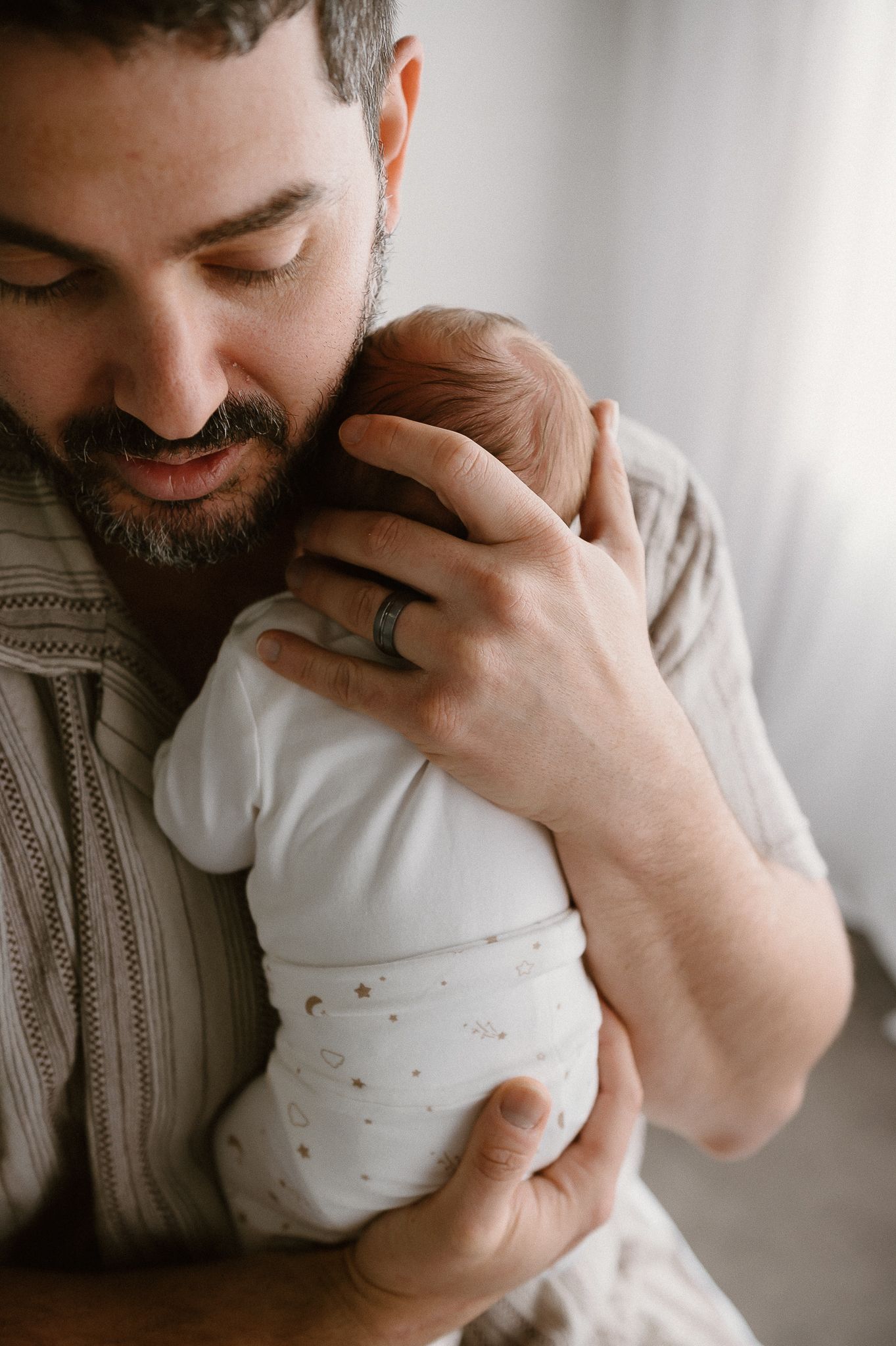 A bearded father tenderly holds his newborn baby against his chest in studio newborn photoshoot with Story&Soul Photography Mandurah WA