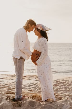 A pregnant couple in white outfits embraces romantically on a sandy beach at sunset with ocean waves for Mandurah Photographer Story & Soul