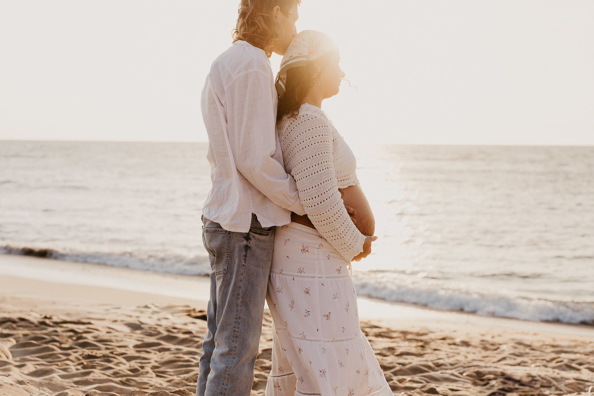 Romantic couple embraces on a sandy beach at sunset with ocean waves in the background holding baby bump. Maternity Photographer Mandurah