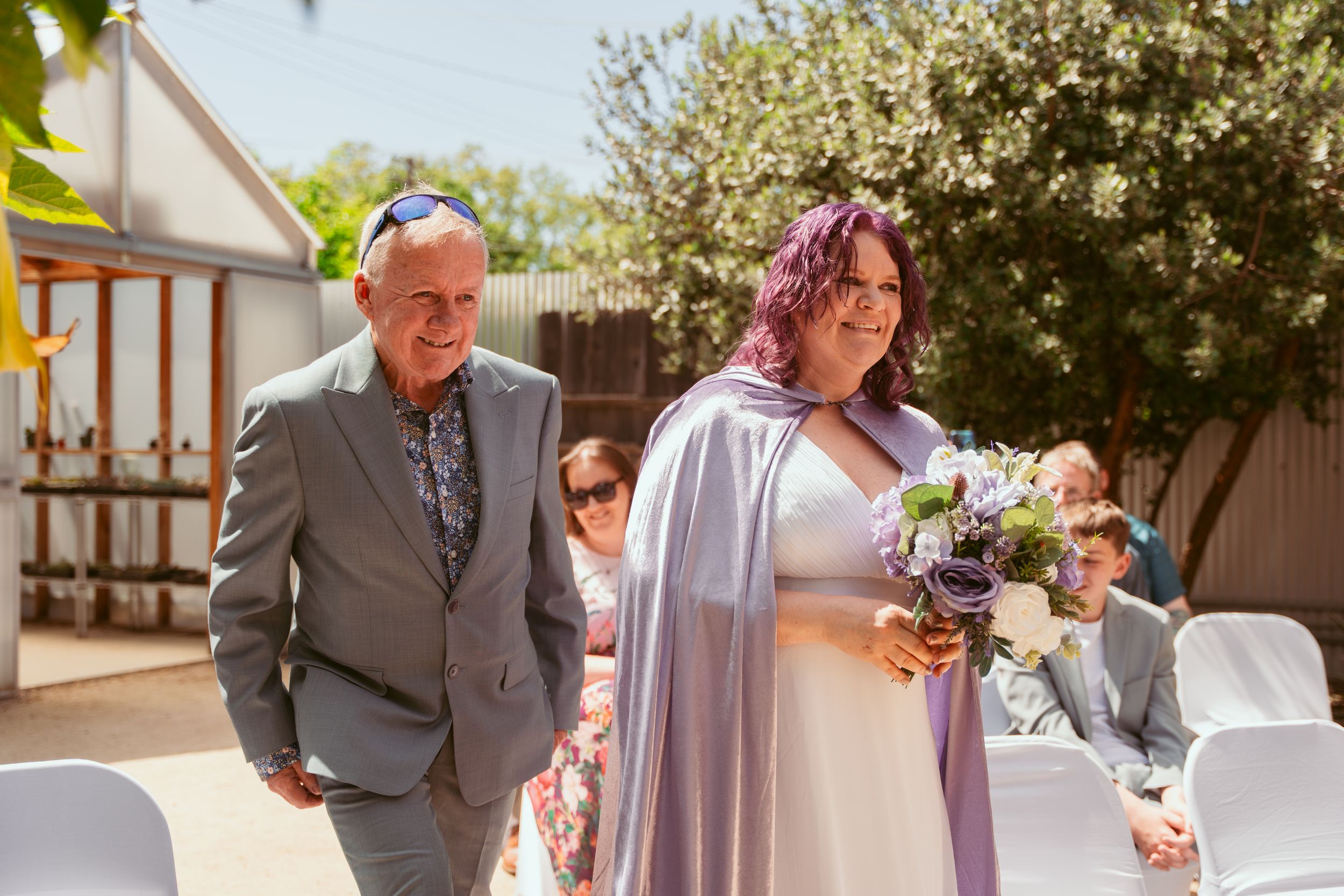 A person in a flowing lavender dress walks down the aisle holding a purple and white bouquet during an outdoor wedding ceremony.