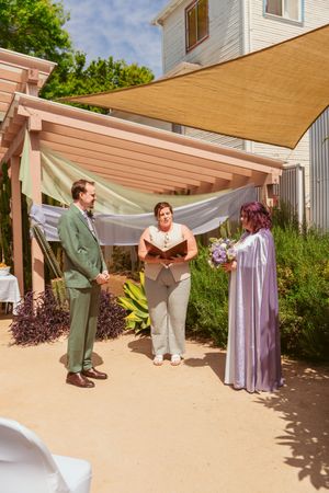 An intimate outdoor wedding ceremony takes place under a beige canvas shade and white fabric draping.