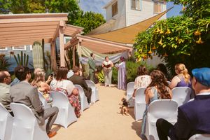 Outdoor wedding ceremony under tan shade sails with white chairs and floral decorations in a garden setting.