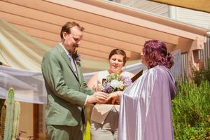 A wedding ceremony takes place outdoors under a pergola with tan sun shade and white fabric decor.
