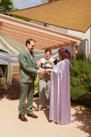 A wedding ceremony takes place in an outdoor garden setting with a tan shade sail overhead.