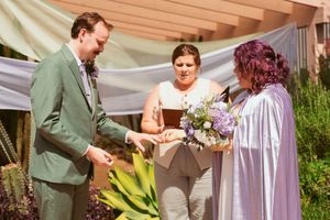 Three people gather at an outdoor ceremony with draped fabric backdrop and purple flowers.